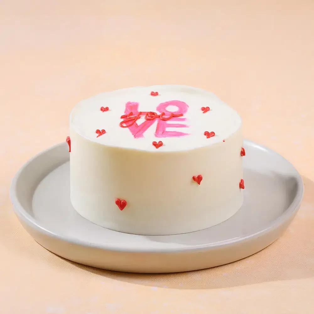 White bento cake with pink palette knife LOVE message and small red icing hearts on a grey plate.