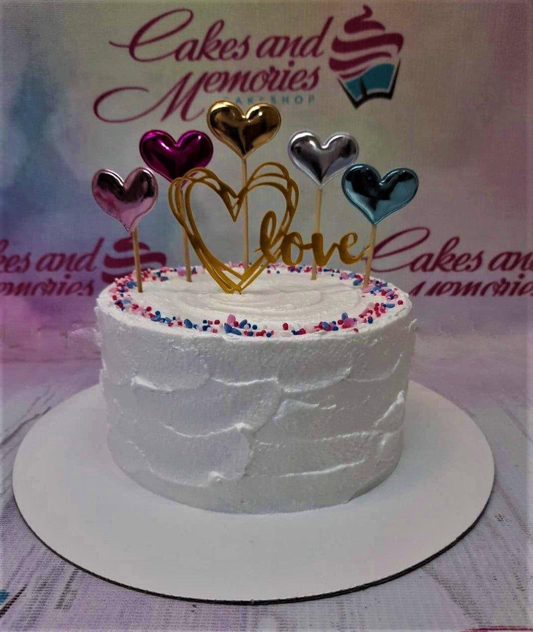 White soft-icing cake with textured spatula finish, colorful sprinkles, and metallic heart toppers with a gold Love sign.