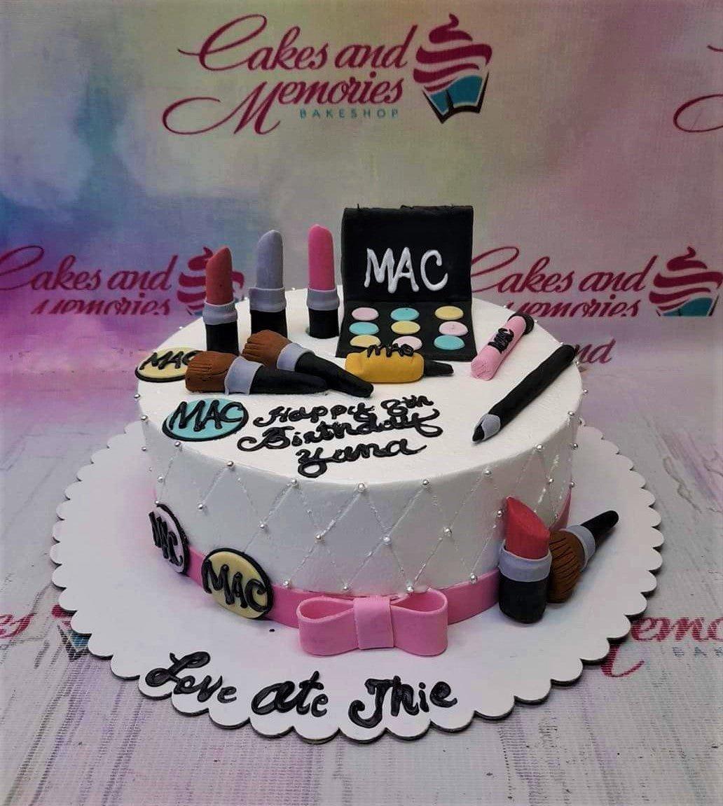 White round cake with MAC makeup fondant toppers including lipsticks, brushes, and eyeshadow palette with pink ribbon and silver pearls.