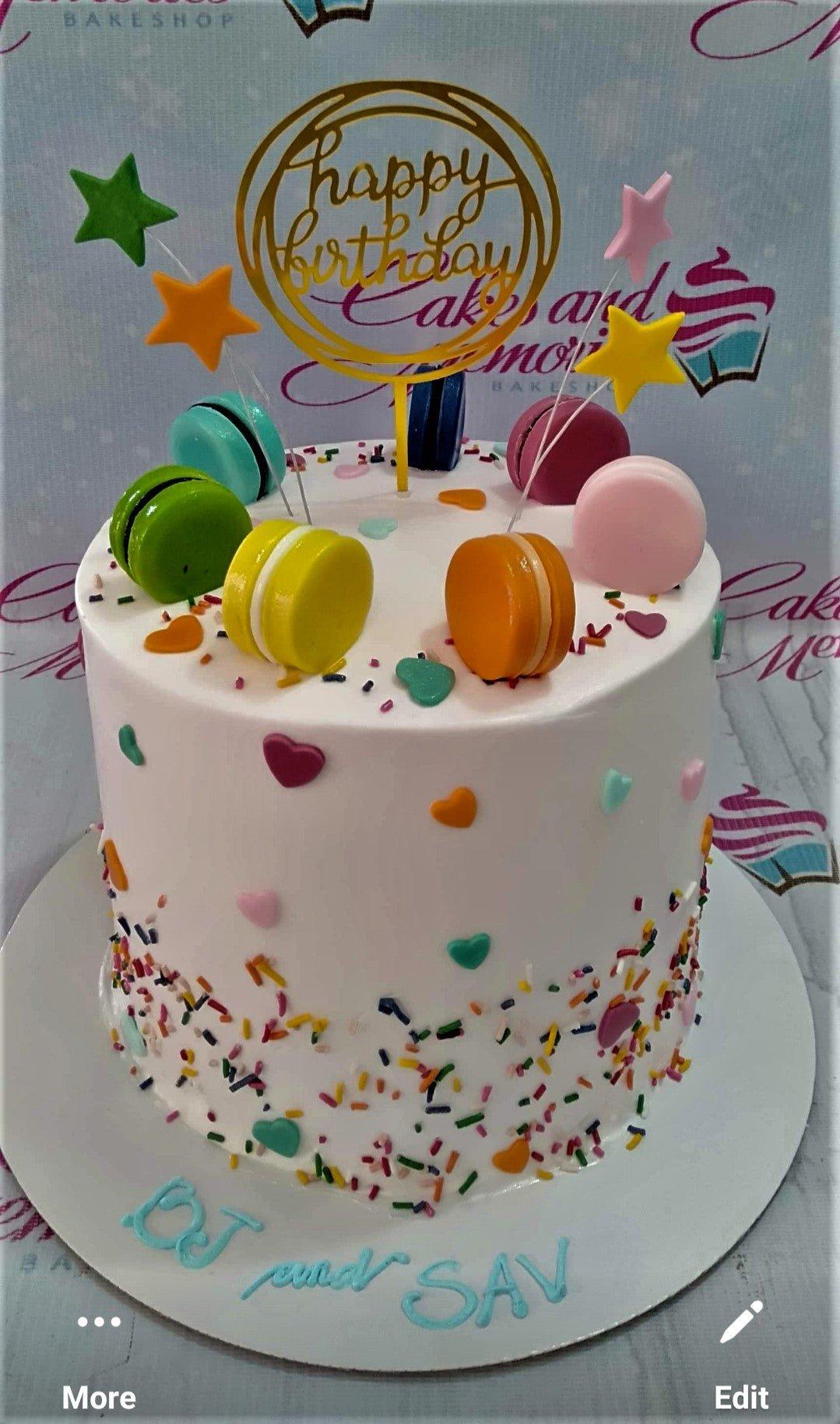 White 1-tier birthday cake with colorful macarons, fondant stars, and a gold Happy Birthday topper.