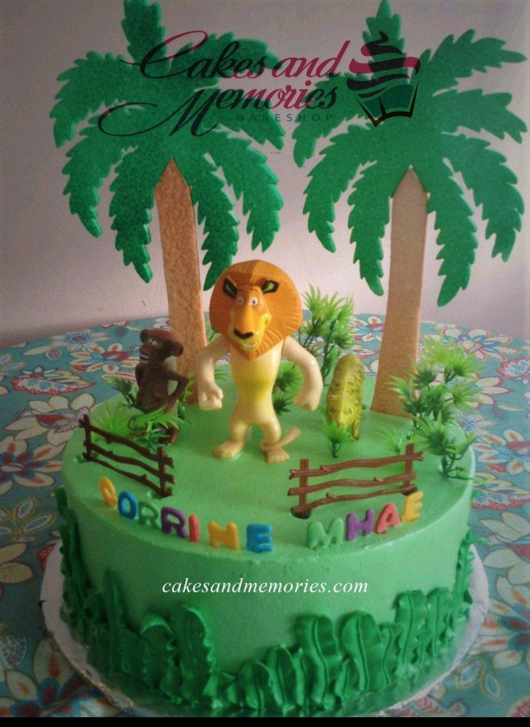 Green jungle themed cake with Madagascar characters Alex the lion and a monkey, featuring palm trees and a fence.
