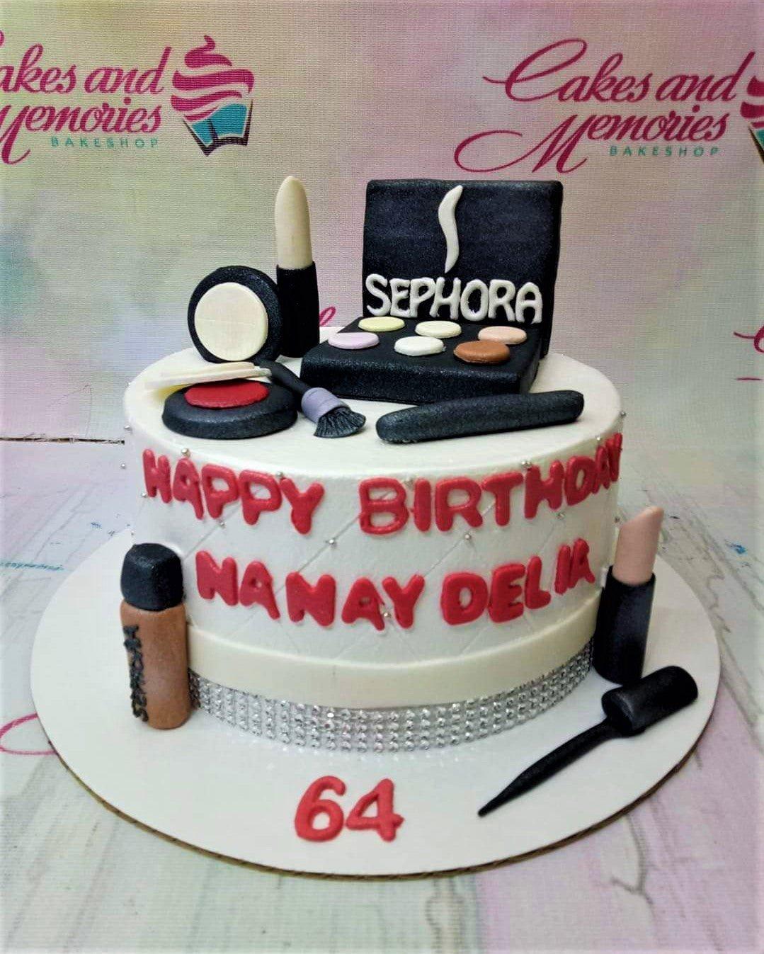 White fondant cake with makeup toppers including Sephora palette, lipsticks, and brushes for a 64th birthday celebration.