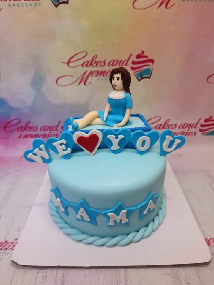 Light blue fondant cake for Mama featuring a sitting woman figurine, blue heart toppers with 'WE LOVE YOU' and 'MAMA' on the side.