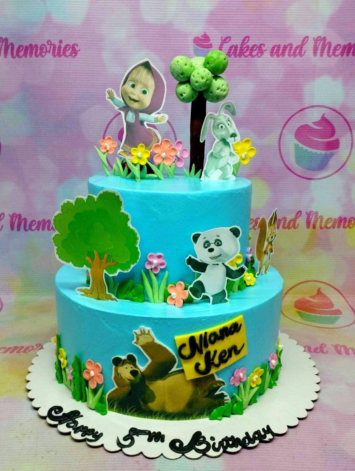 Two-tier light blue Masha and the Bear themed birthday cake with character printouts, fondant trees, and colorful flowers.