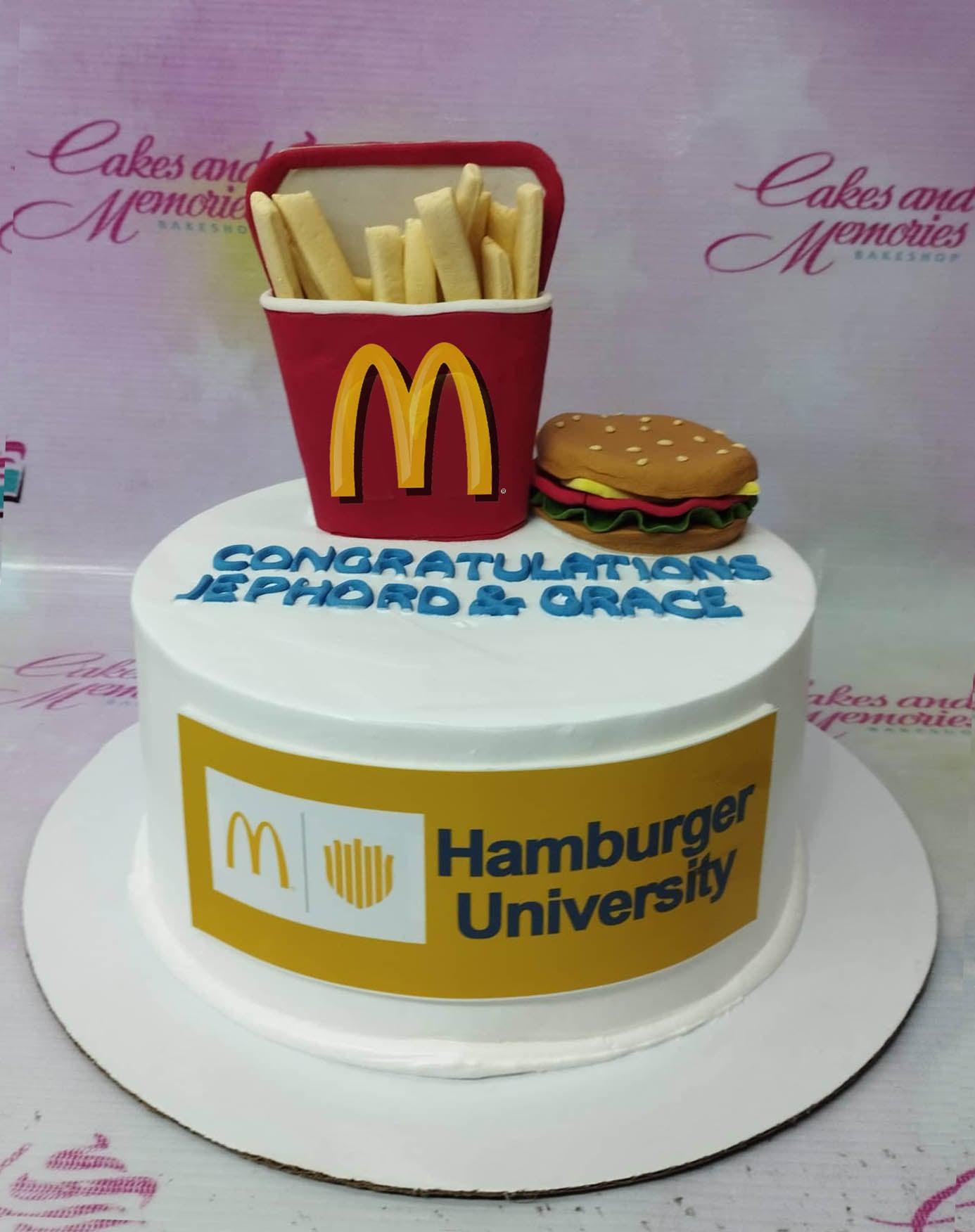 White round cake featuring a 3D fondant McDonald's french fries container and a hamburger topper with a Hamburger University logo on the side.