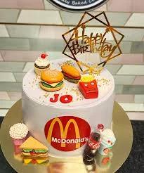 White McDonald's themed birthday cake with fondant burger, fries, and hotdog toppers and a gold Happy Birthday sign.