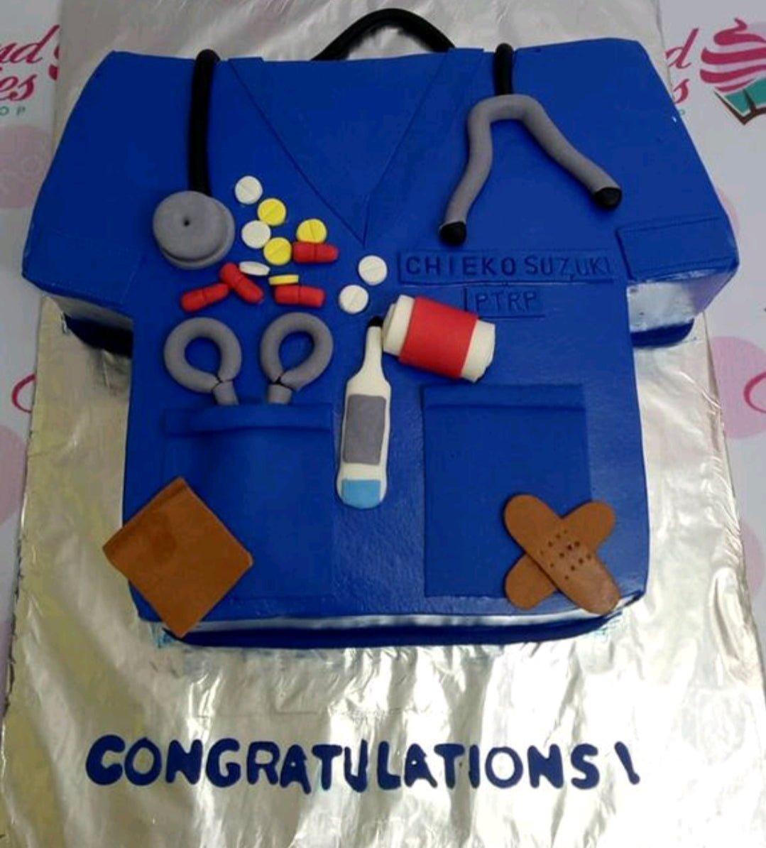 Blue medical scrub shaped cake with fondant stethoscope, thermometer, pills, and bandages for a graduation or professional celebration.