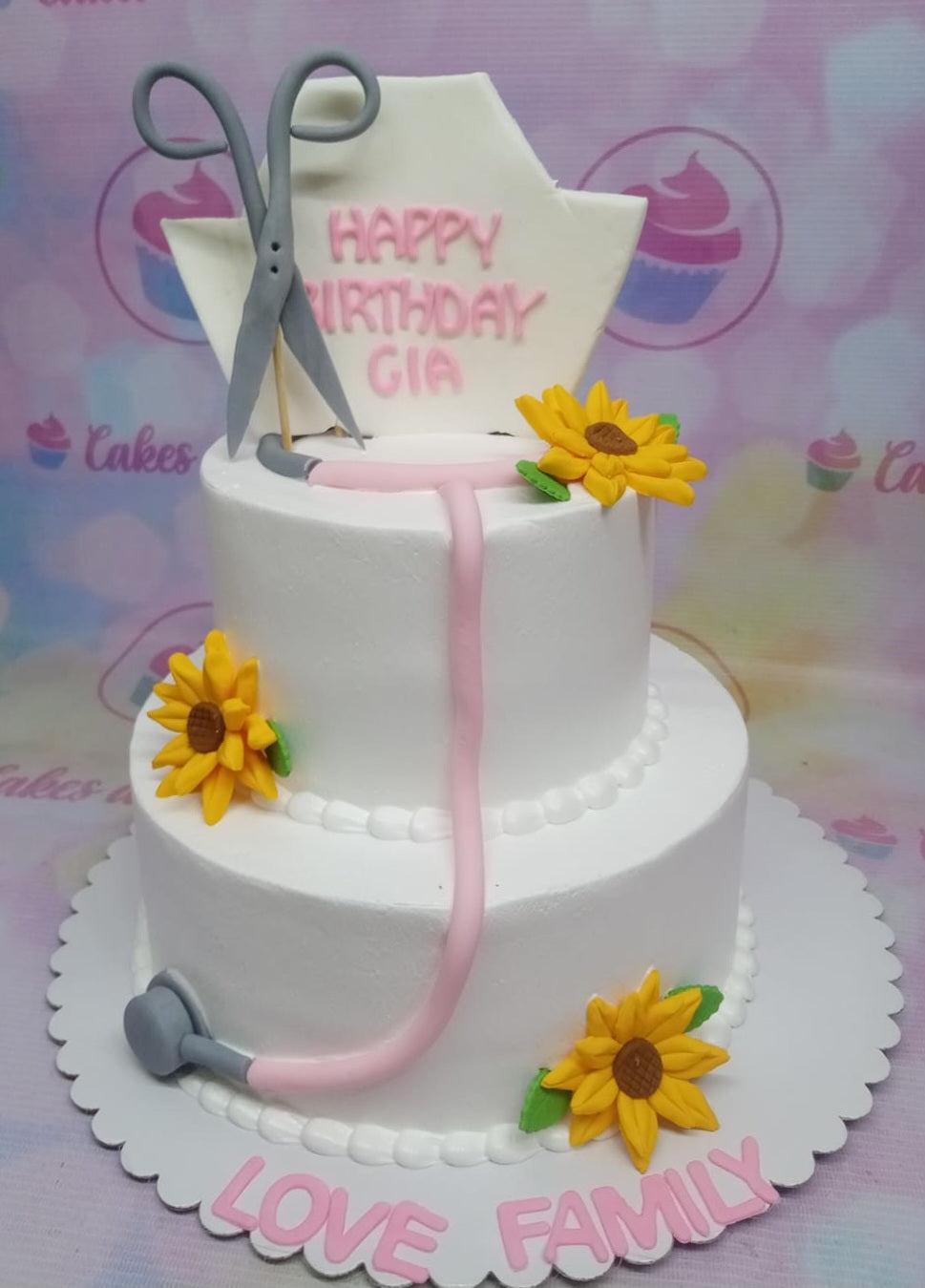 White 2-tier medical themed birthday cake with fondant sunflowers, a pink stethoscope, and grey scissors.