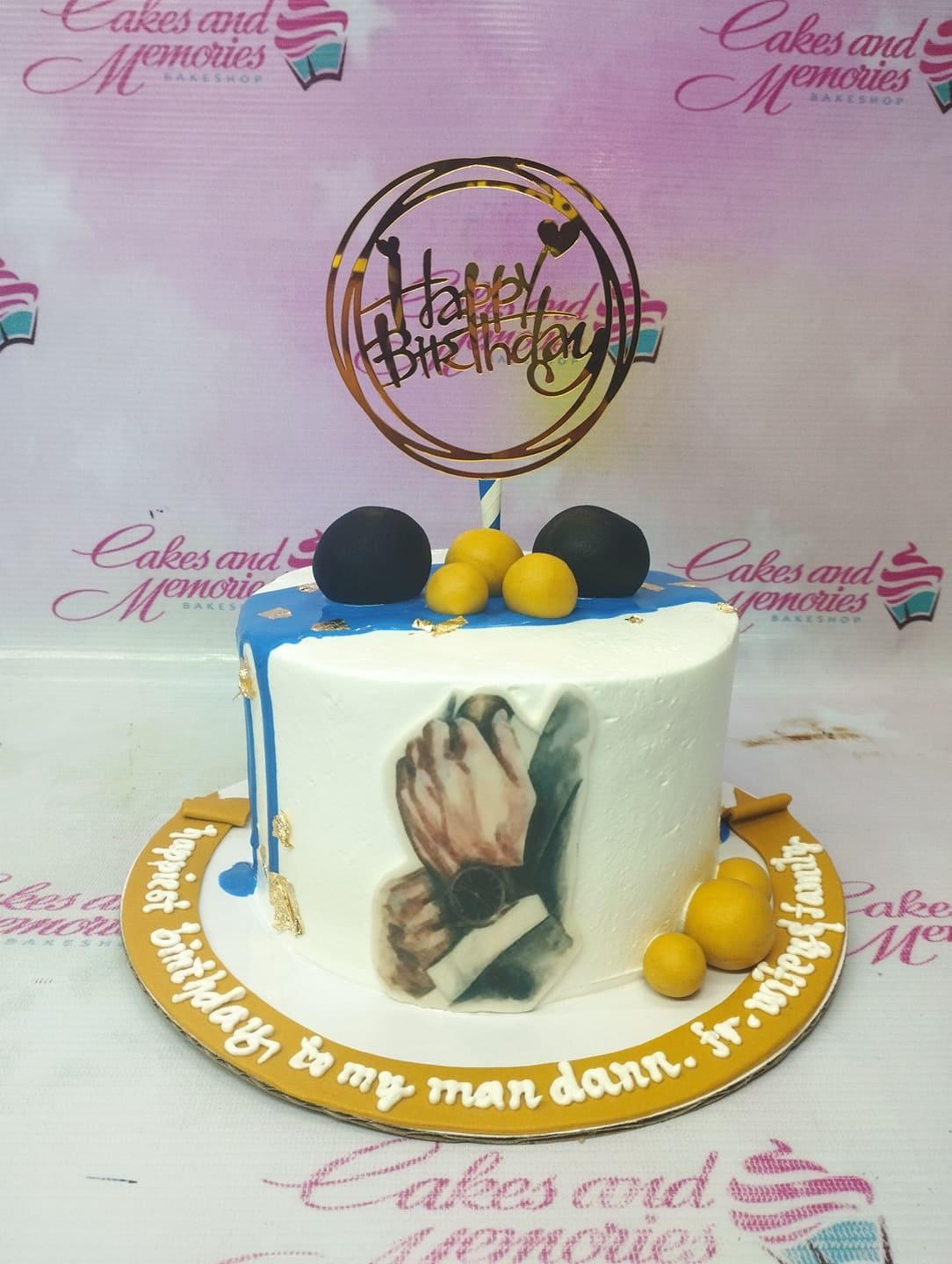 White minimalist cake with blue drip, gold leaf accents, and a photo print of hands with a watch.