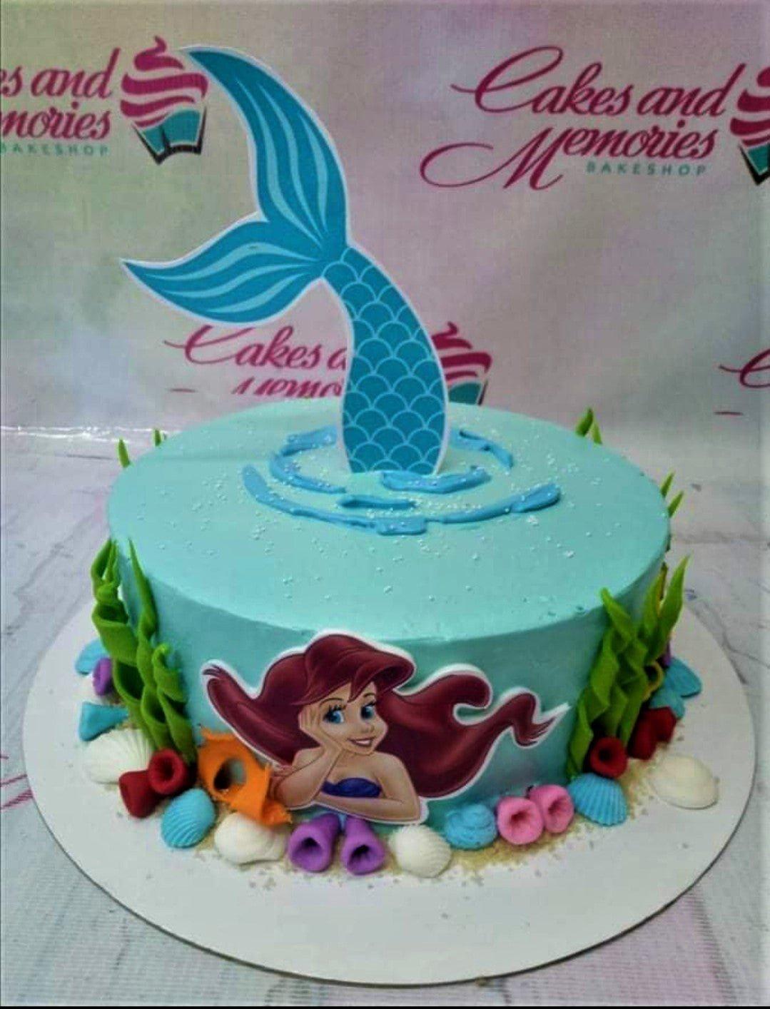 Light blue mermaid themed cake with a large blue mermaid tail topper and a printed Ariel character on the side surrounded by fondant sea life.