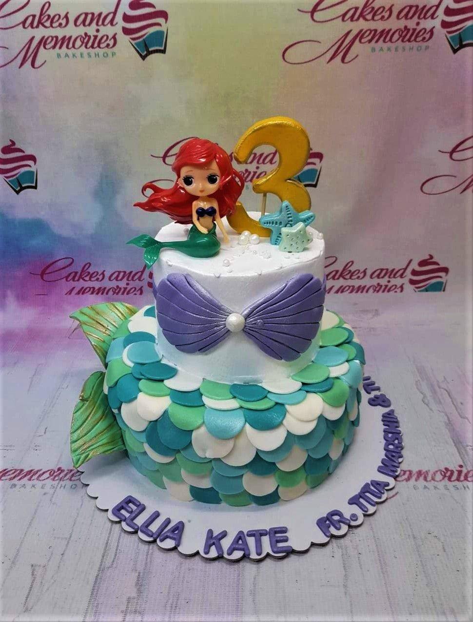 Two-tier mermaid themed cake with white icing, teal and green scales, a large purple bow, and a Little Mermaid Ariel figurine on top.