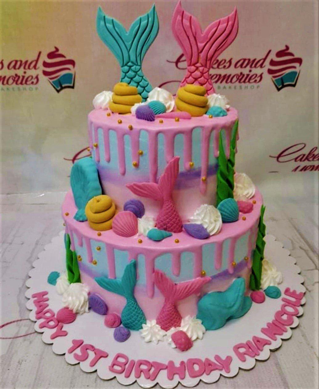 Two-tier mermaid themed birthday cake in pink and blue with fondant mermaid tails, shells, and seaweed decorations.