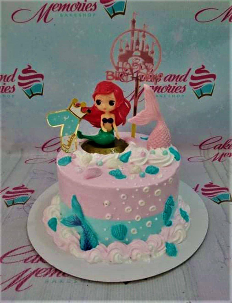 Pink and light blue mermaid themed birthday cake with a red-haired mermaid figurine, pink mermaid tail, and seashell icing decorations.