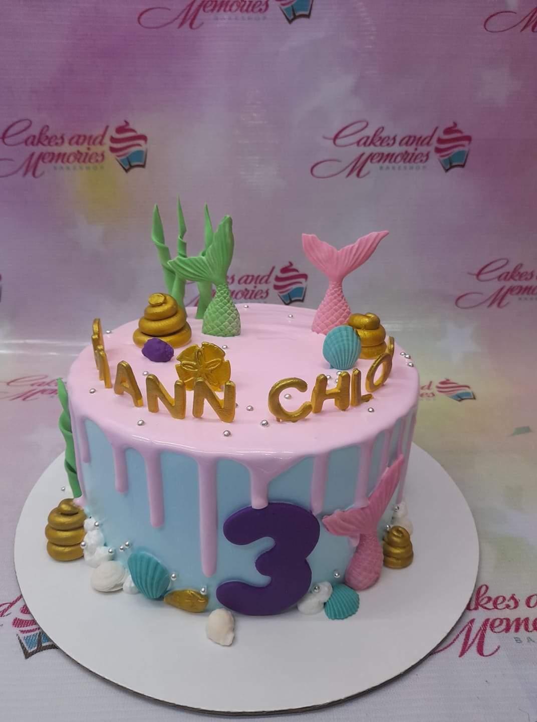 Light blue mermaid themed cake with pink drip, mermaid tails, shells, and gold name toppers.