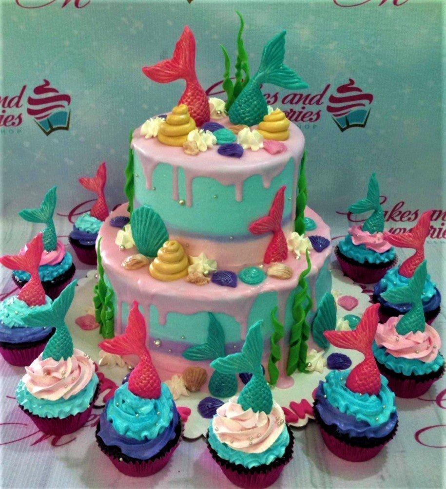 Two-tier mermaid themed cake with light blue and pink icing, decorated with colorful mermaid tails, seashells, and seaweed, surrounded by matching cupcakes.