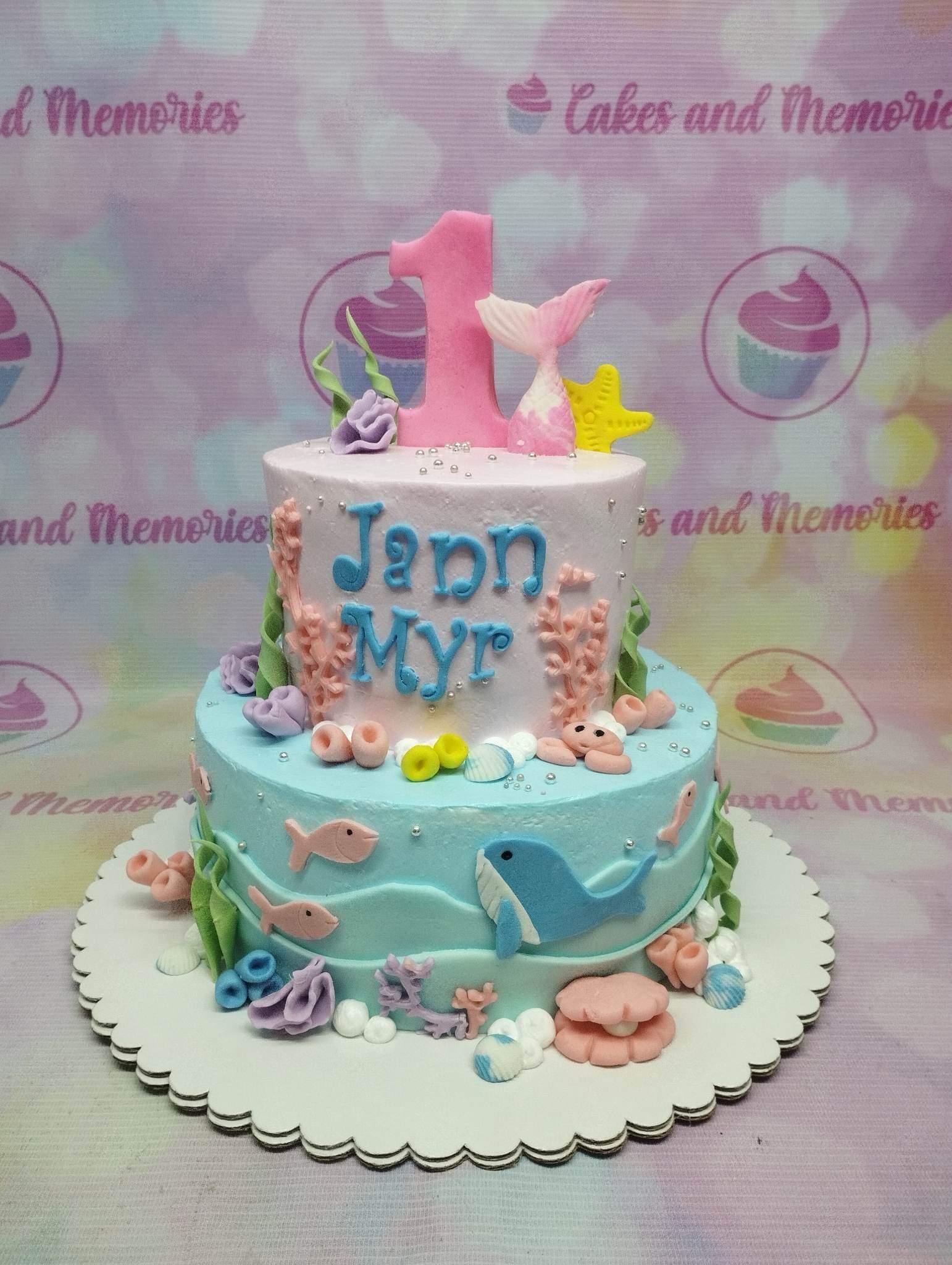 Two-tier under the sea themed cake in pink and light blue with a mermaid tail, number one topper, and fondant sea creatures.