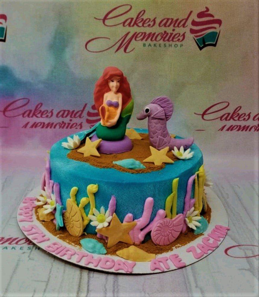 Under the sea themed blue cake with a fondant mermaid and seahorse topper, decorated with colorful coral and starfish.