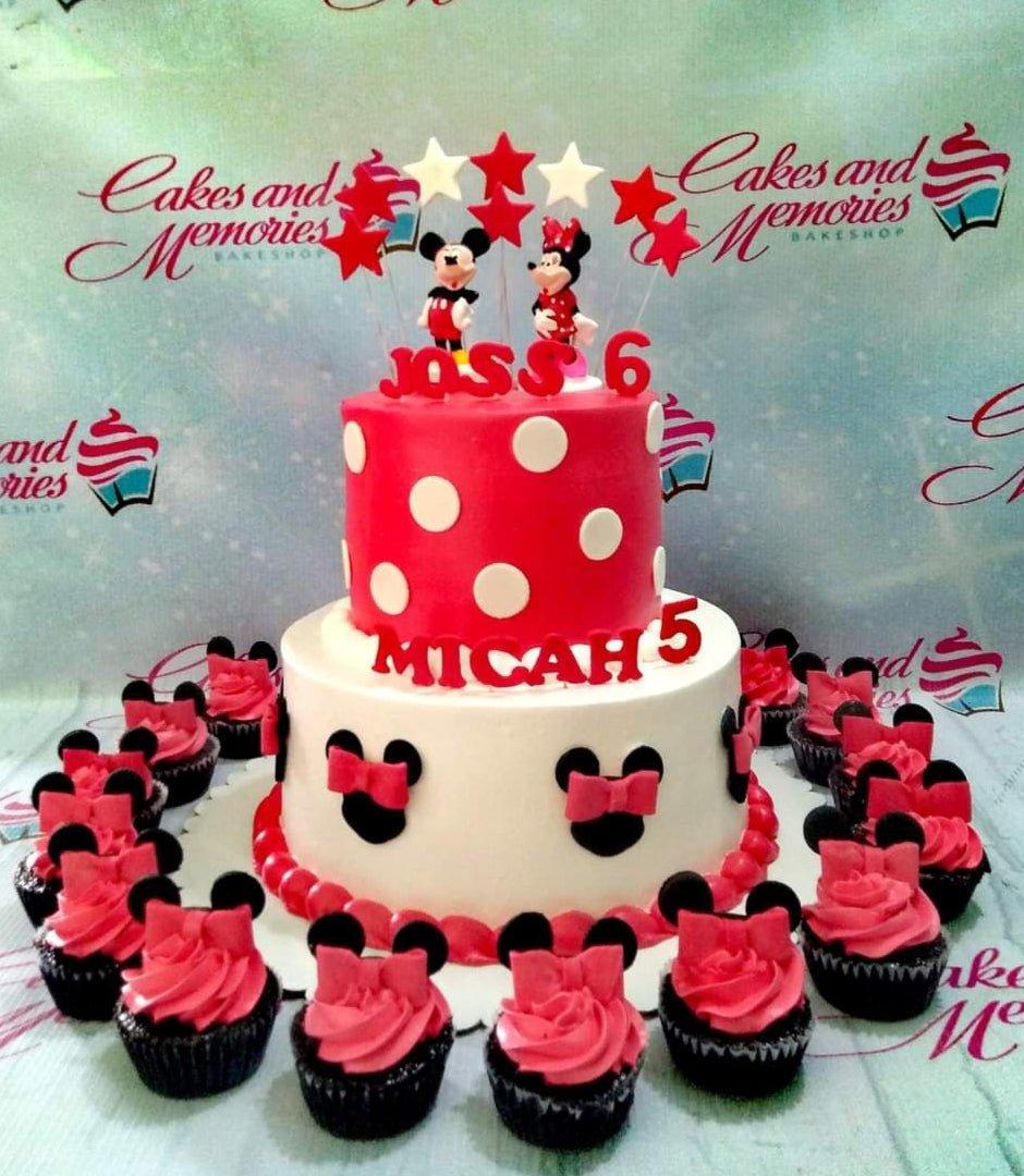 Two-tier Mickey and Minnie Mouse themed cake with red polka dots, black mouse silhouettes, and pink bows, surrounded by matching cupcakes.