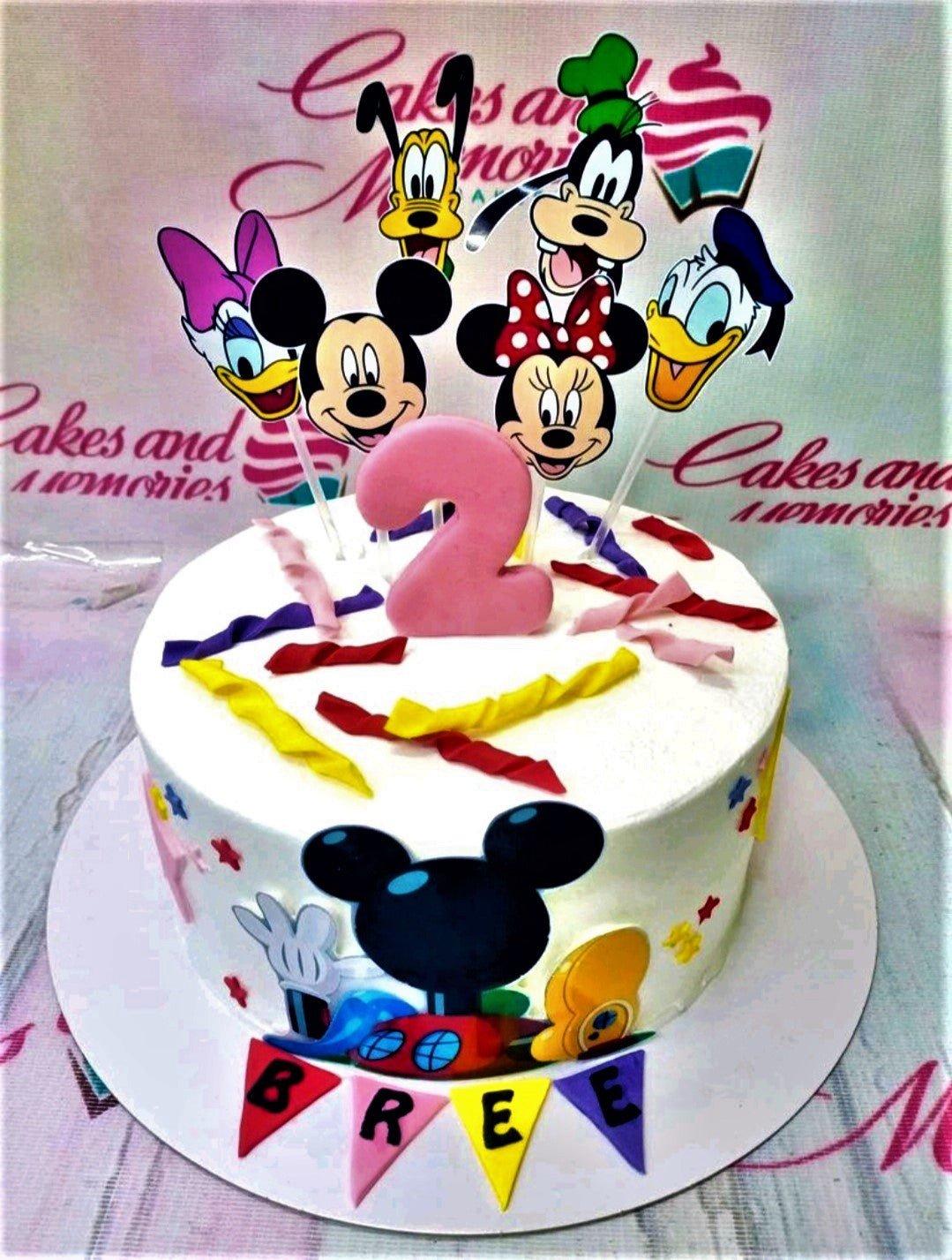 White Mickey Mouse and Friends themed birthday cake with colorful fondant streamers and character printouts.