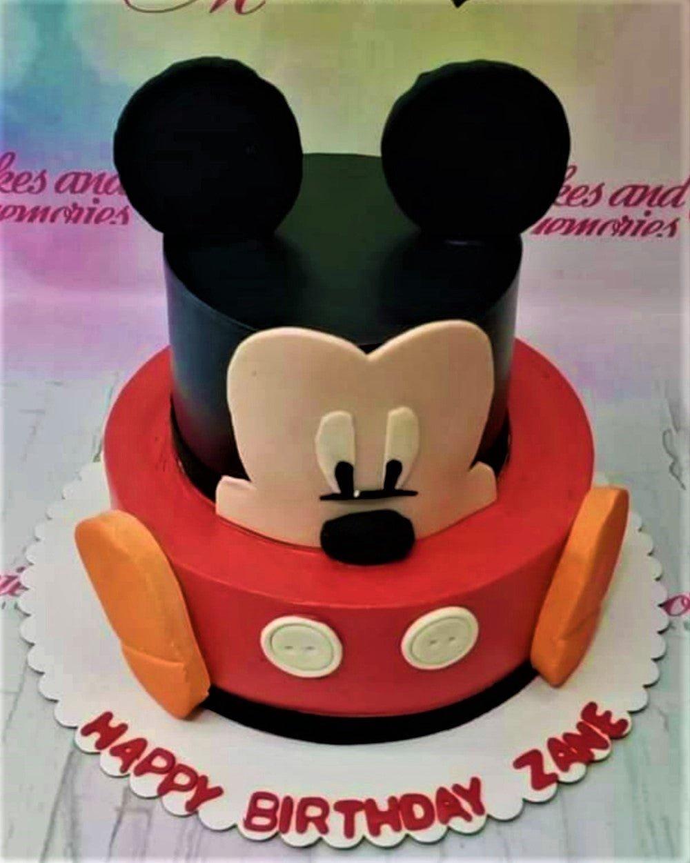 Two-tier Mickey Mouse themed fondant cake with a black top tier featuring ears and a red bottom tier with yellow shoes and white buttons.