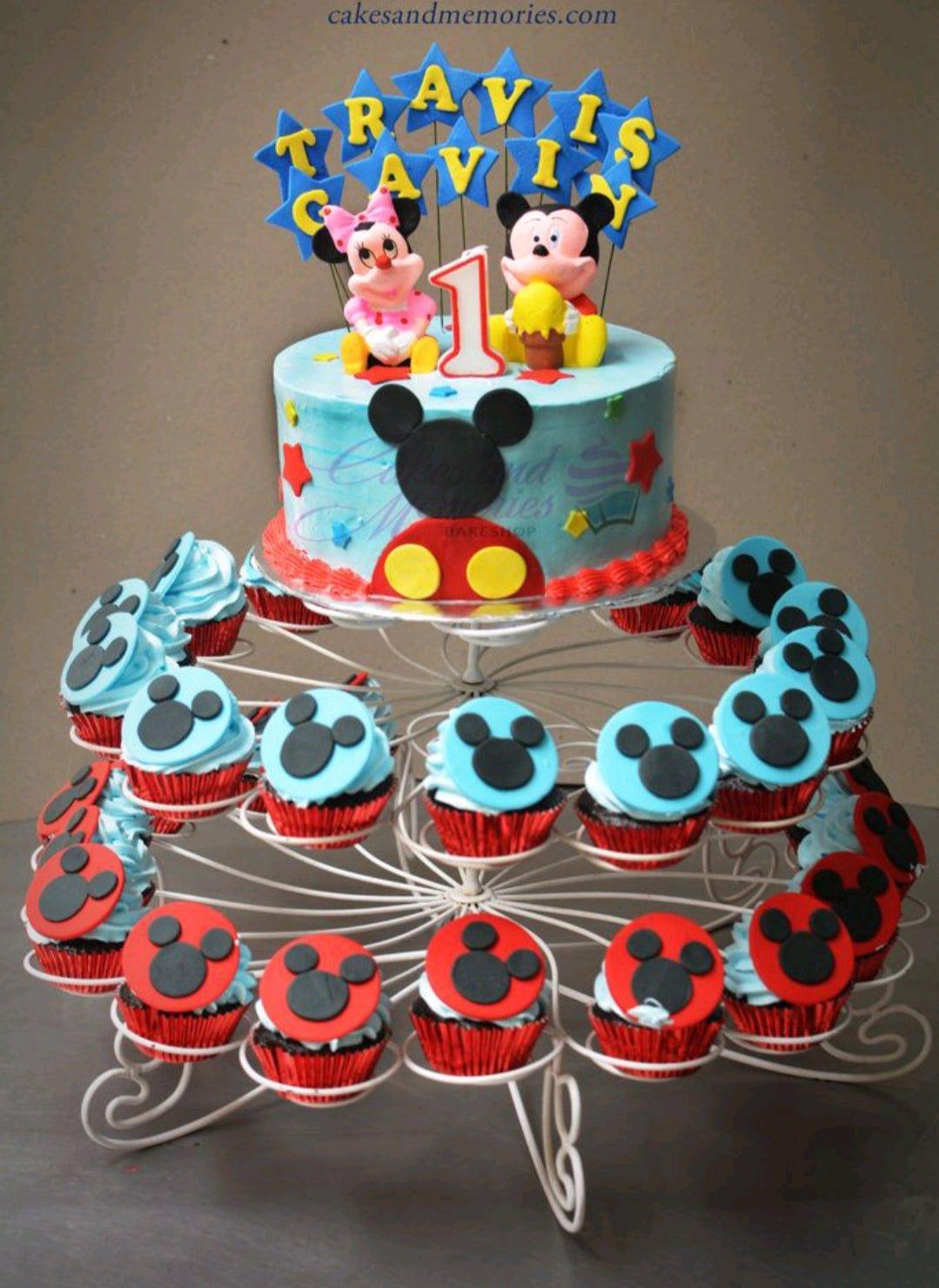 Mickey and Minnie Mouse themed 1st birthday cake with blue icing, red stars, and character toppers on a white cupcake stand.