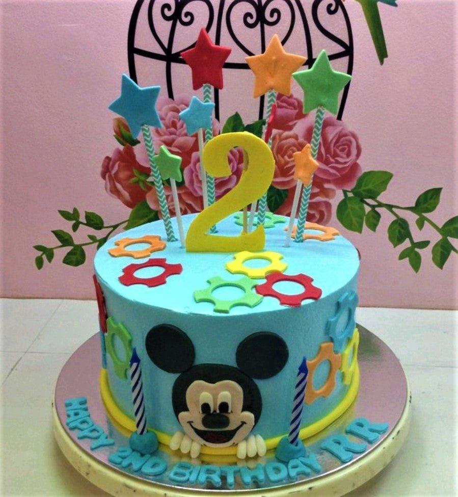Light blue Mickey Mouse birthday cake with colorful fondant gears, stars on sticks, and a large yellow number 2 topper.