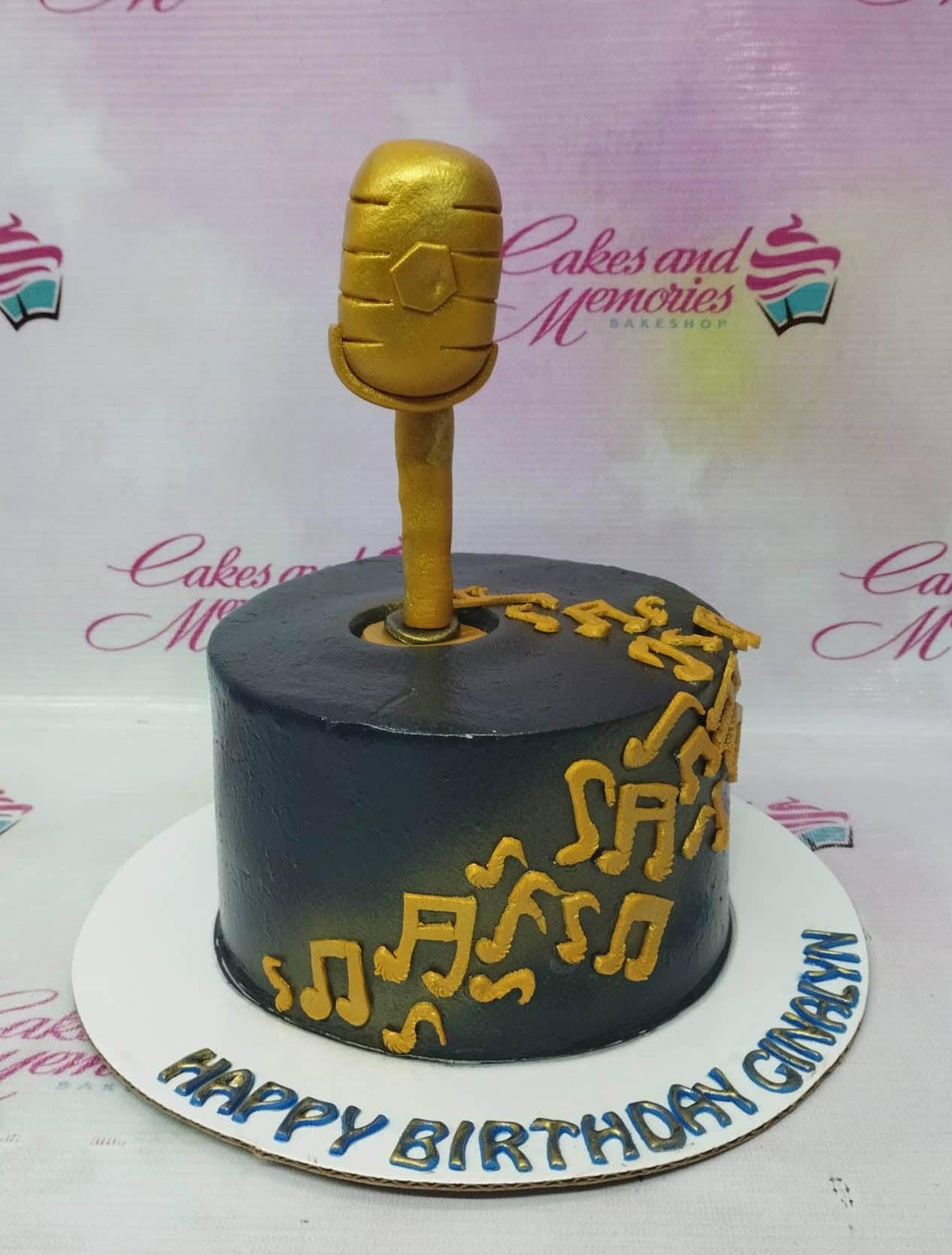 Black round cake with a large gold 3D microphone topper and gold musical notes on the side.