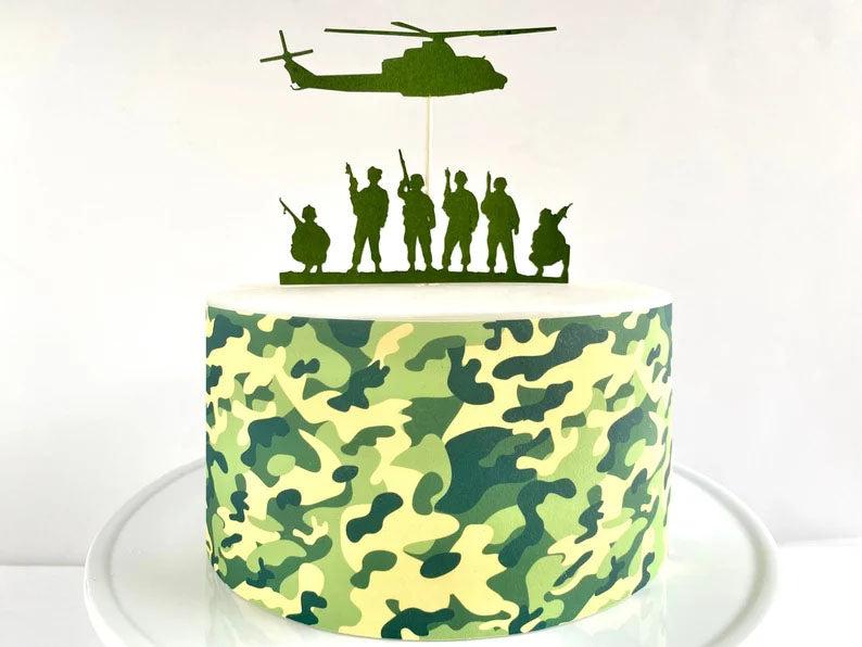 Military themed camouflage cake with green helicopter and soldier silhouette toppers on a white background.