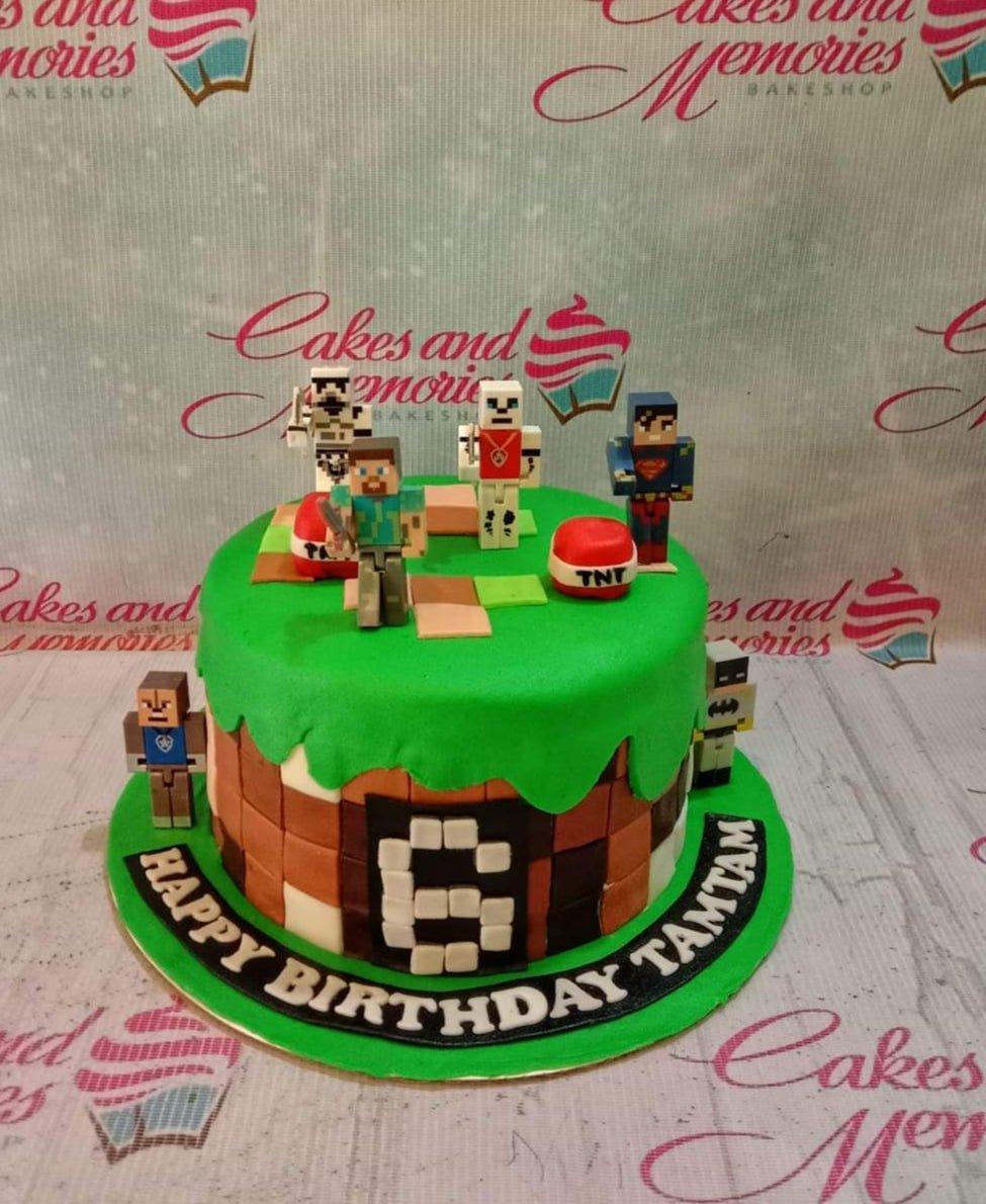 Minecraft themed birthday cake with Steve, Superman, and Batman figures on a green grass and brown pixelated base.