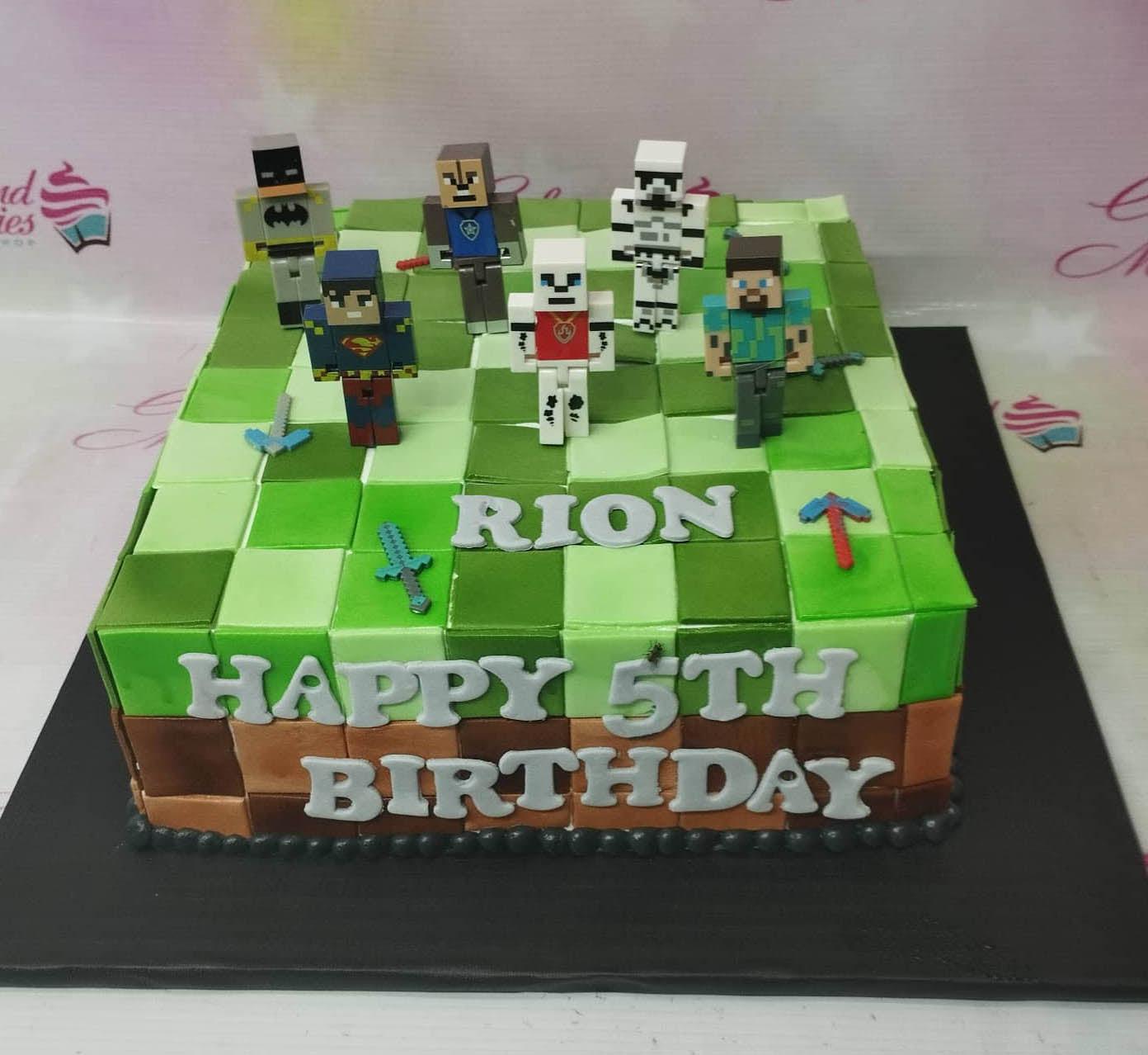 Minecraft themed square fondant cake with pixelated green grass pattern and 3D character figures including Steve and Superman.