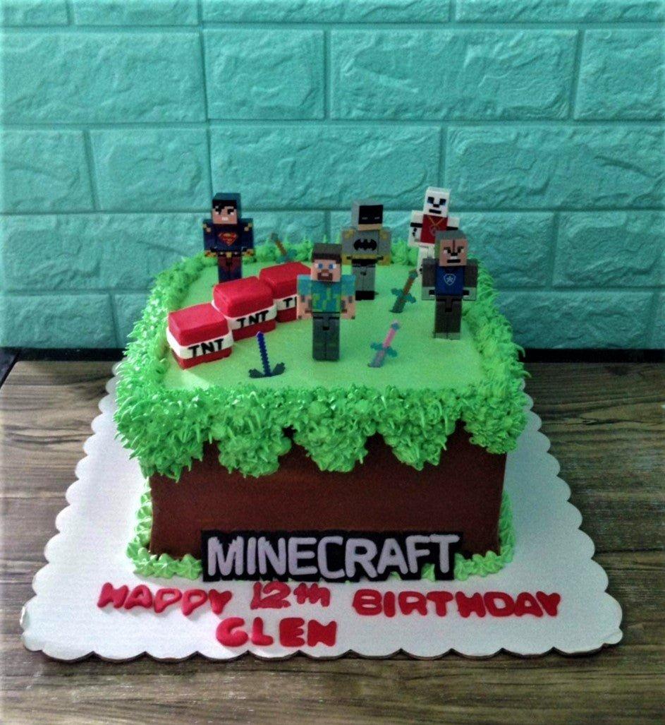 Minecraft themed square cake with brown icing, green grass piping, TNT blocks, and character figurines including Steve, Batman, and Superman.