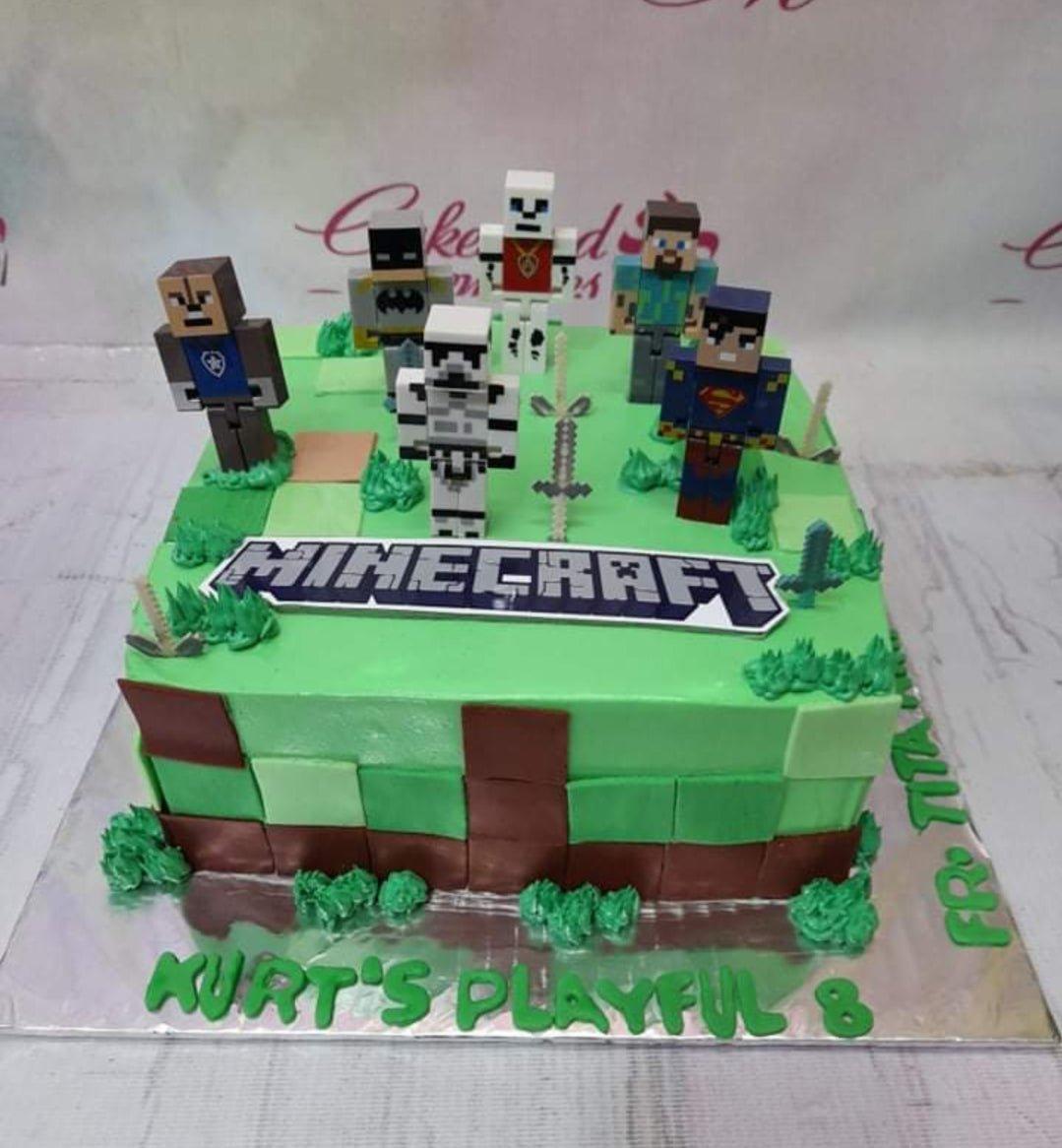 Square Minecraft themed birthday cake with green grass icing, pixelated side panels, and various character figurines on top.
