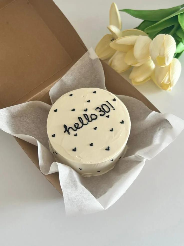 White bento cake with black piped icing hearts and 'hello 30!' message in a brown box with white tissue paper.
