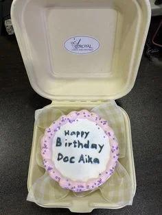 White bento cake with pink icing borders and purple sprinkles featuring a Happy Birthday Doc Aika message.