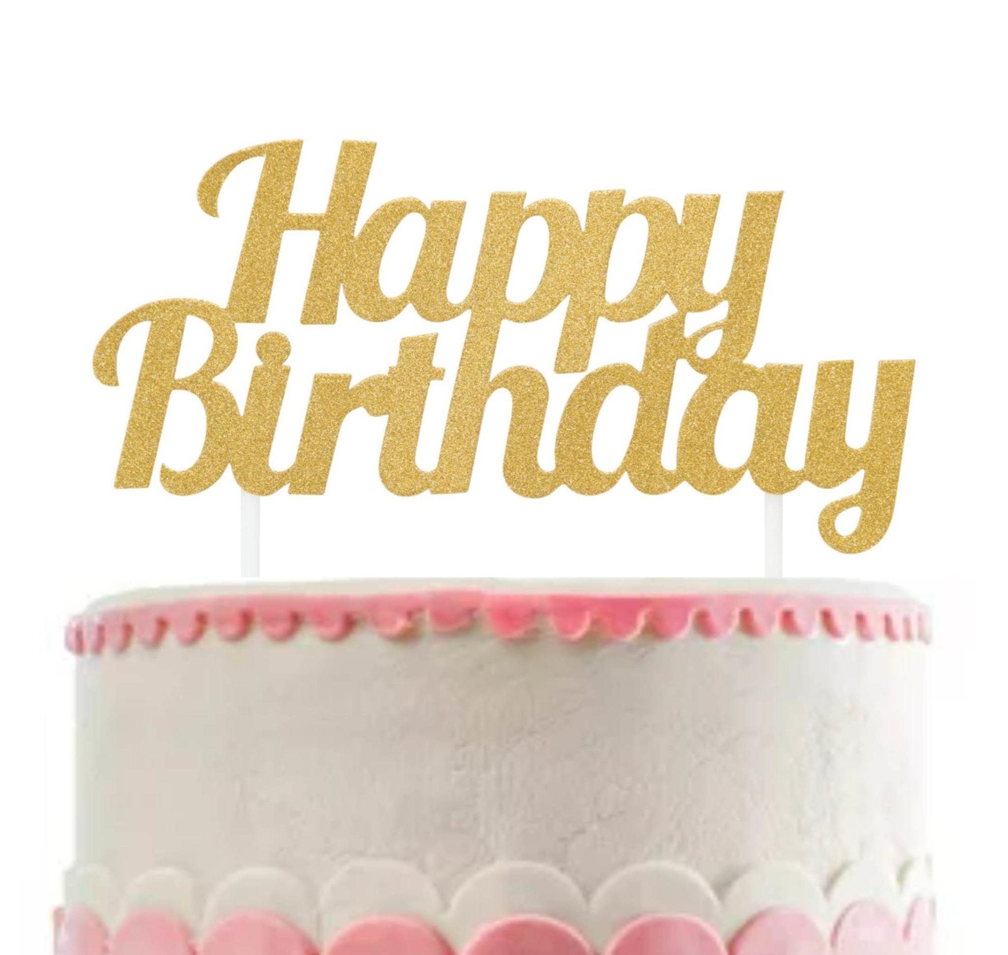 White minimalist birthday cake with pink icing scallops and a large gold glitter Happy Birthday topper.