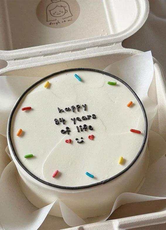 Minimalist white bento cake with rainbow sprinkles and a black icing message reading happy 34 years of life.