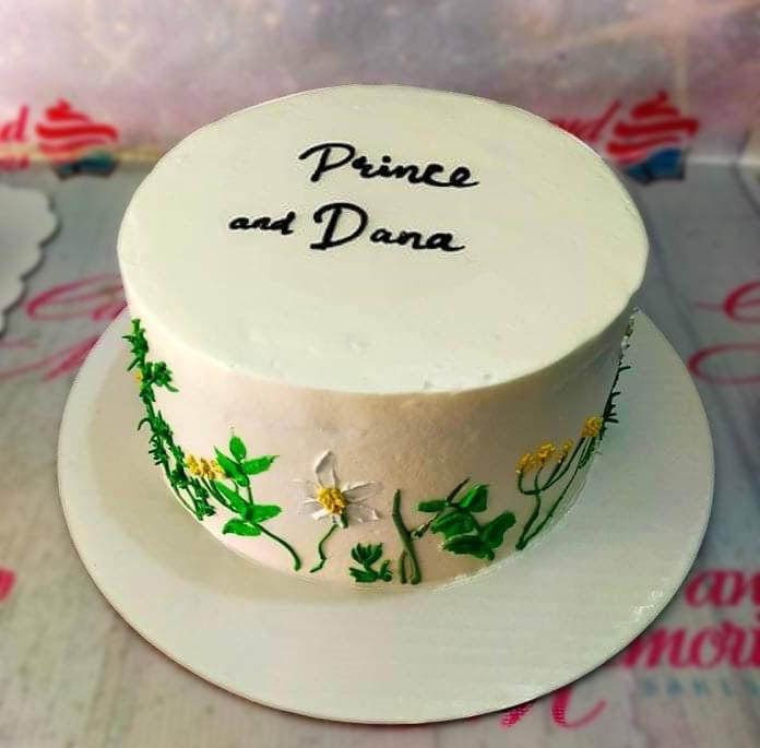 Minimalist white cake with hand-painted icing flowers and green stems on the side, featuring black script text on top.