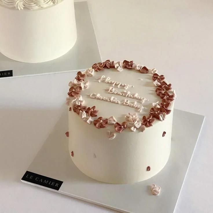 Minimalist white 1-tier birthday cake with a ring of small pink and brown piped icing flowers and a handwritten message.
