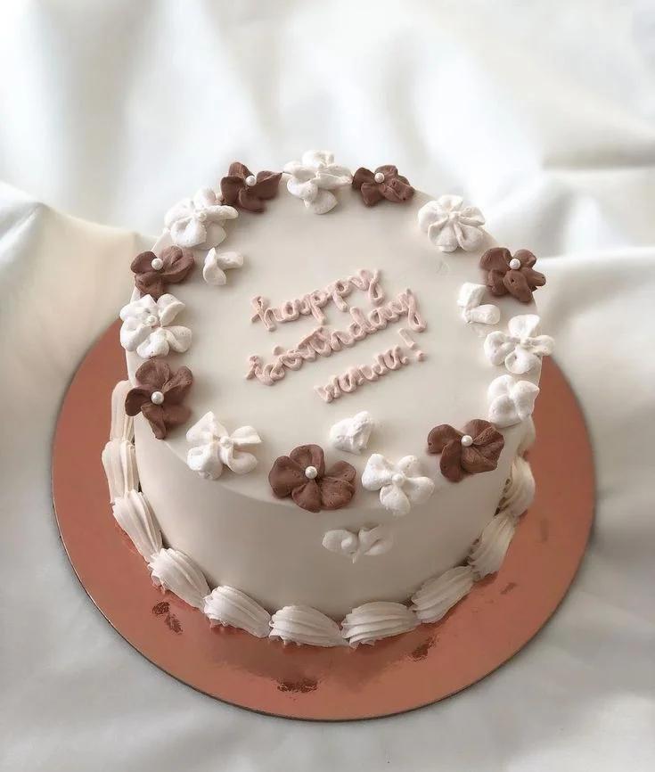Minimalist white birthday cake with brown and white piped icing flowers and a pink 'Happy Birthday' message.