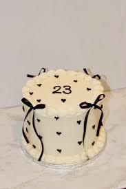 White minimalist birthday cake with black piped hearts, black ribbon bows, and the number 23 on top.