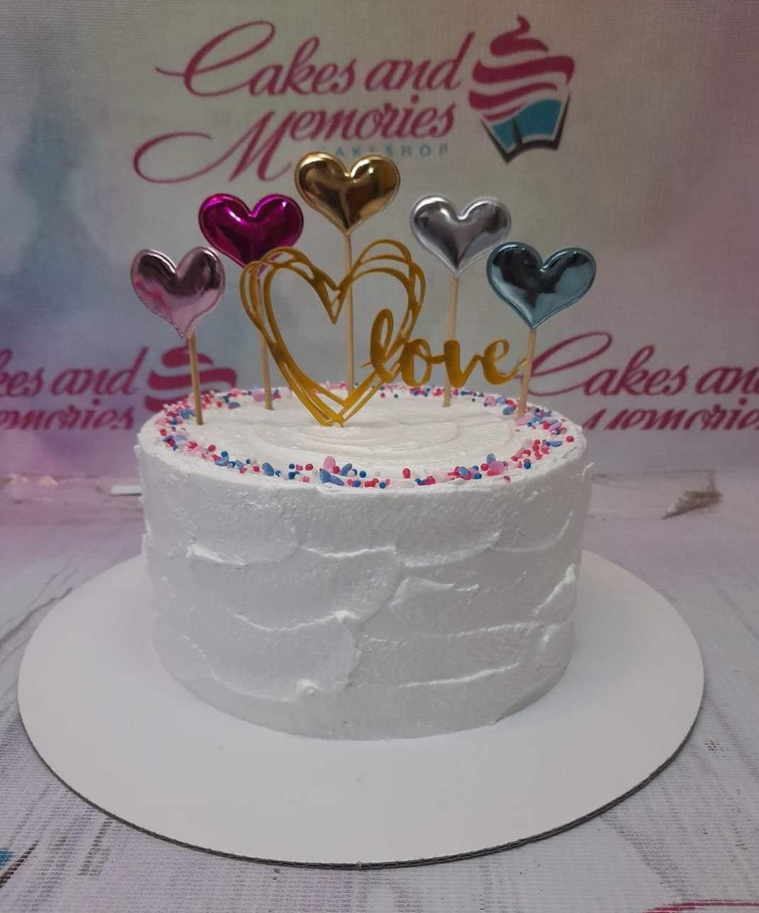 White minimalist cake with textured icing, colorful sprinkles, and metallic heart toppers in gold, silver, pink, and blue.