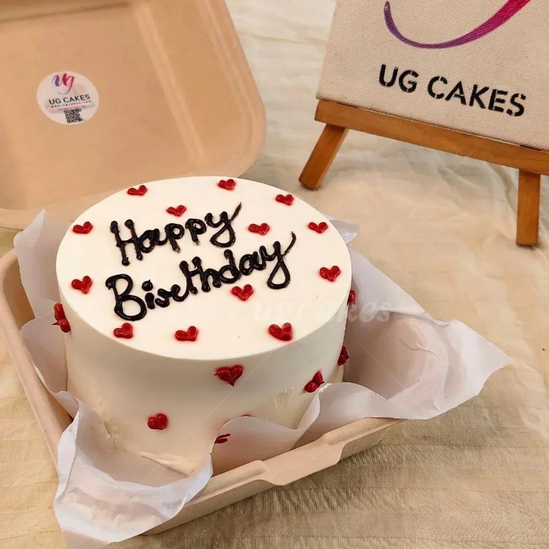 White bento cake with red icing hearts and black piped Happy Birthday message in a tan takeout box.