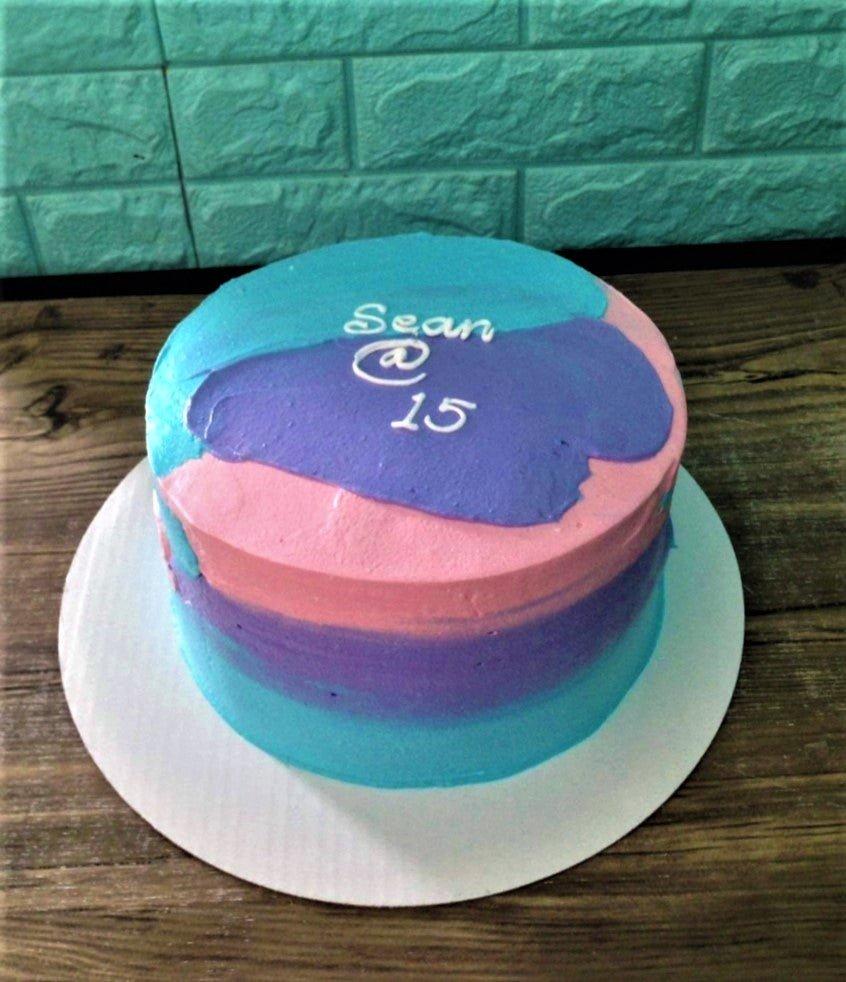 A minimalist round cake with a colorful palette knife icing design in blue, pink, and purple with a white piped message.