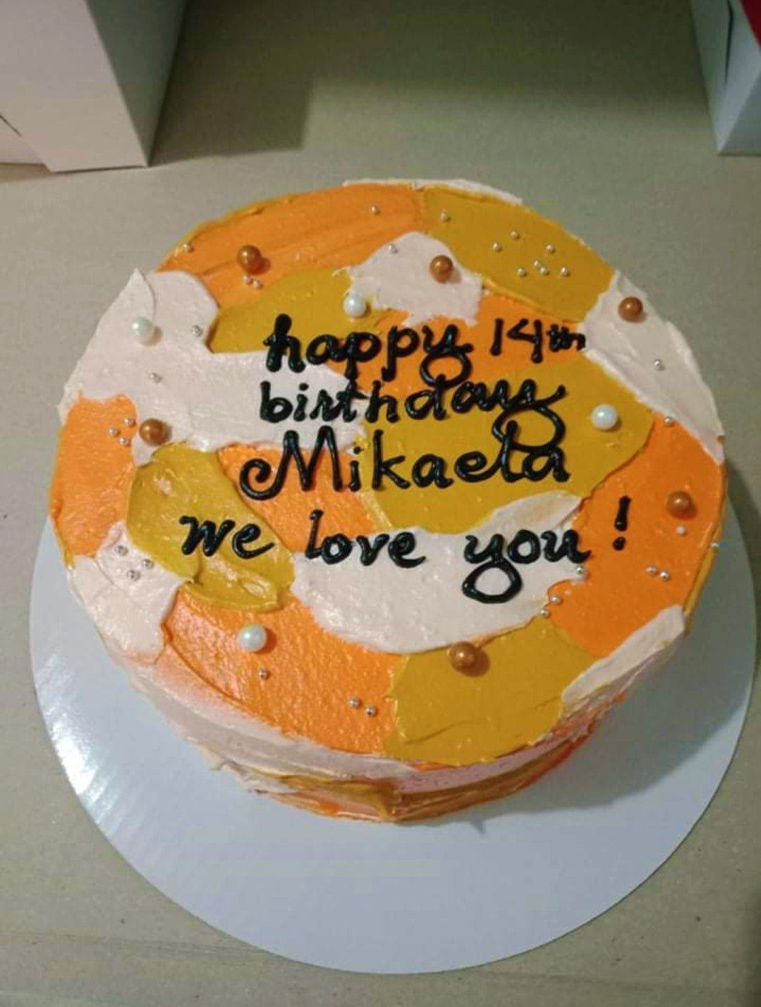 Round minimalist birthday cake with orange, yellow, and white palette knife icing textures, gold and white pearls, and black piped message.
