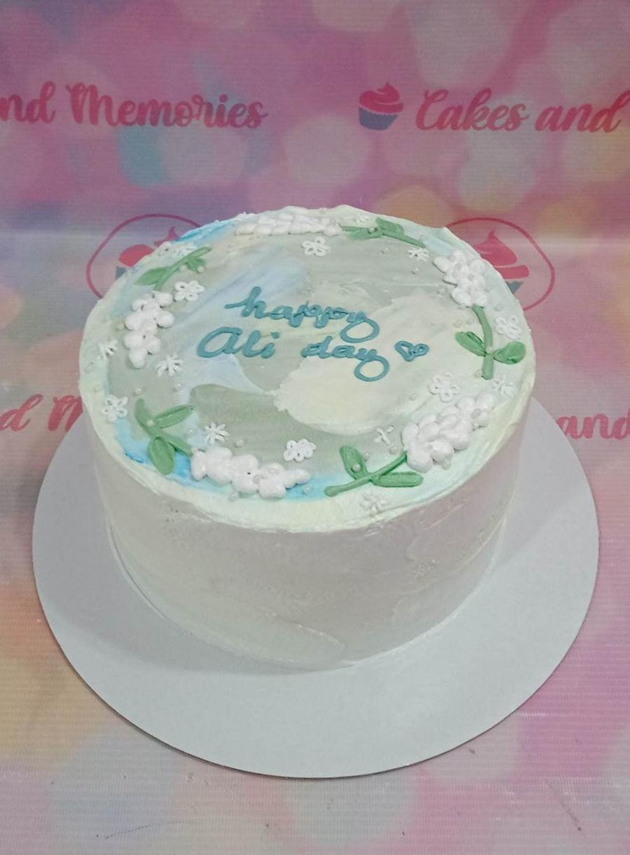 Minimalist white bento-style cake with blue and green palette knife icing, white piped flowers, and 'happy Ali day' message.