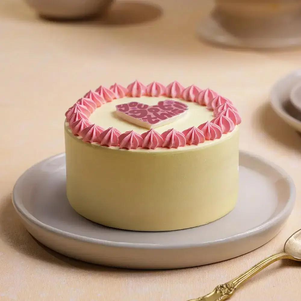 Minimalist light yellow bento cake with pink icing rosettes and a heart-shaped 'You Got This' message topper.