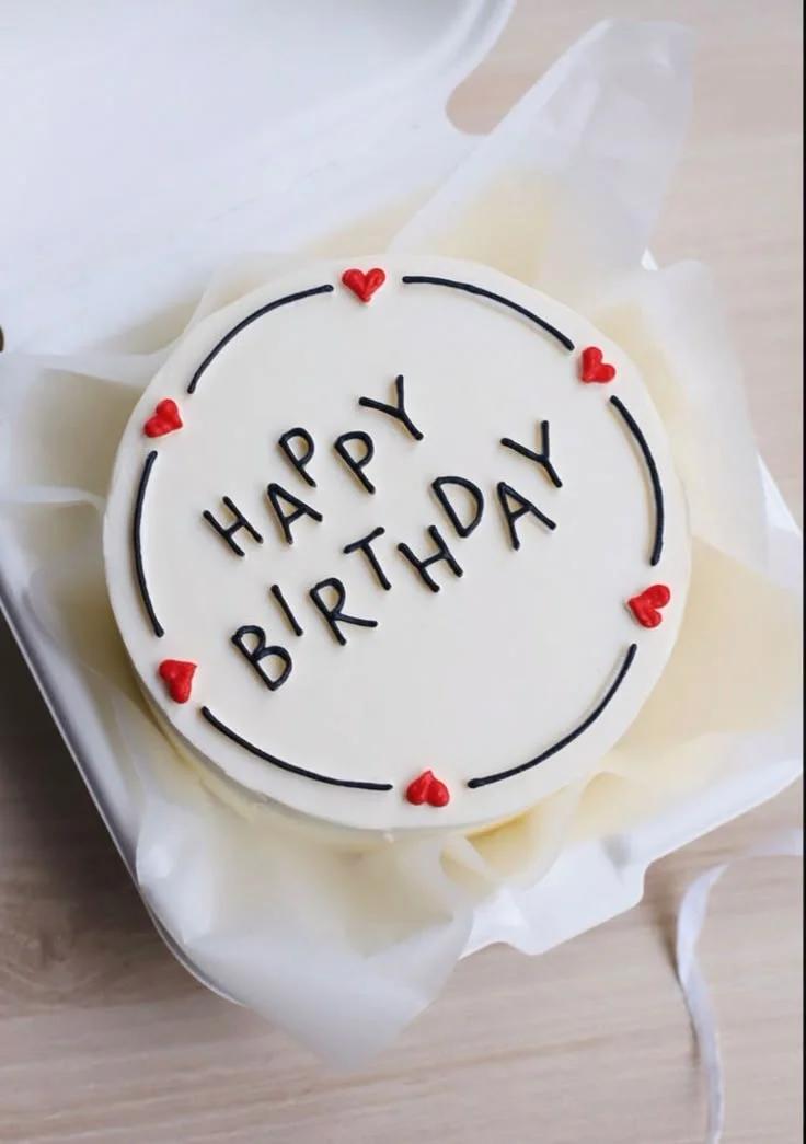 White bento cake with black piped 'HAPPY BIRTHDAY' text and small red icing hearts inside a black circular border.