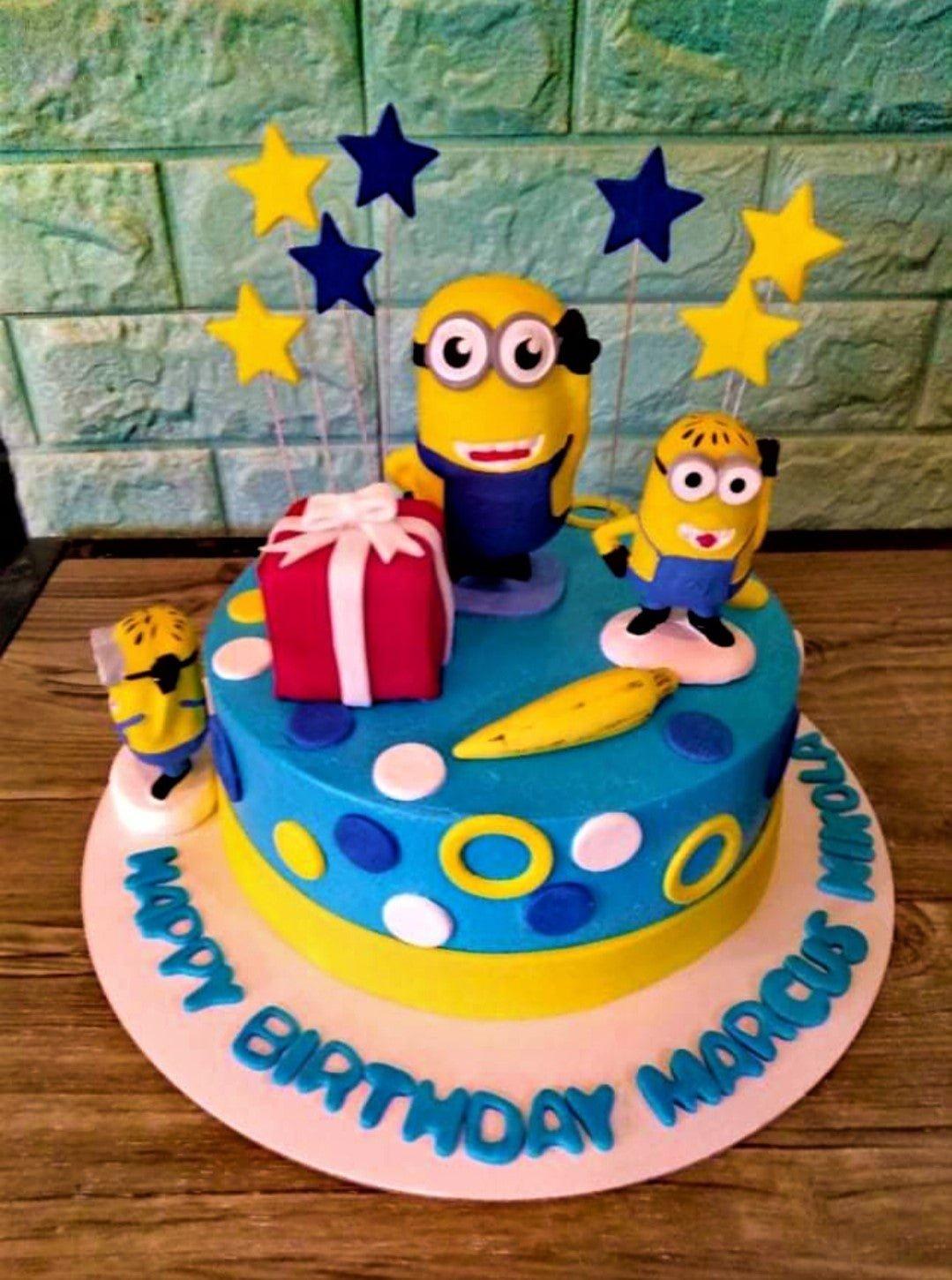 Minions themed birthday cake with yellow and blue fondant, featuring 3D Minion figures, a red gift box, and blue and yellow stars.