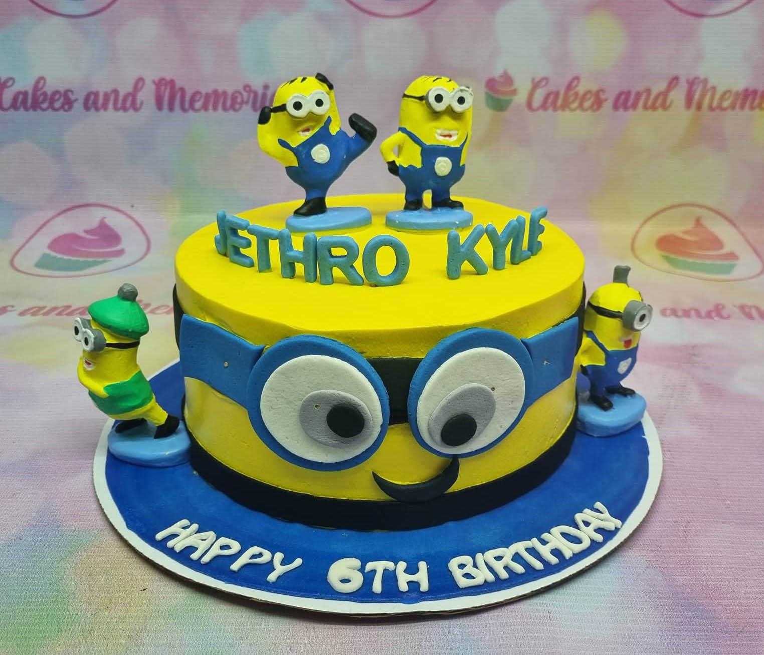 Yellow Minions themed birthday cake with 3D fondant figures, blue gumpaste letters, and a large Minion eye design on the side.