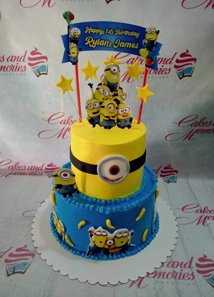 Two-tier Minions themed birthday cake with yellow and blue icing, featuring various Minion character printouts and yellow stars.